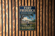 A vintage metal sign for Fort Frederick National Monument in Georgia, featuring a scenic illustration of the monument surrounded by a moat and a bridge.