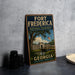A vintage-style metal sign for Fort Frederick National Monument in Georgia, featuring a scenic illustration of the monument surrounded by a tree and a bridge.