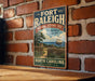 A vintage-style metal sign for the Fort Raleigh National Historic Site in North Carolina, featuring a ship and a wooden fence, mounted on a brick wall.