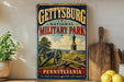 A vintage-style metal sign for Gettysburg National Military Park in Pennsylvania, featuring a cannon and a statue, is displayed on a shelf next to a wooden cutting board and some lemons.