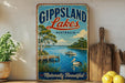 A vintage-style sign for Gippsland Lakes, Australia, featuring a scenic image of a lake with a boat and a bird, surrounded by lush greenery and a dock.
