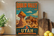 A vintage-style metal sign for Goblin Valley State Park, featuring a desert landscape with rock formations and mountains, is displayed on a shelf next to a wooden cutting board and some lemons.