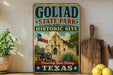 A vintage-style metal sign for Goliad State Park and Historic Site, featuring an illustration of the historic building and a cannon, with the text "Preserving Texas History" and "GOLIAD STATE PARK AND HISTORIC SITE".