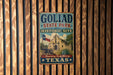 A vintage-style metal sign for Goliad State Park and Historic Site, featuring a historical building and a Texas flag, is displayed on a wooden wall.
