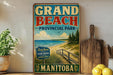 A vintage-style sign for Grand Beach Provincial Park in Manitoba, featuring a beach scene with a wooden walkway leading to the sand and water.