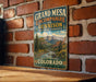 A vintage-style metal sign for Grand Mesa, Colorado, featuring a scenic mountain landscape with a river and pine trees.