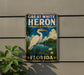 A vintage-style metal sign for the Great White Heron National Wildlife Refuge in Florida, featuring a large white heron standing on a pond surrounded by palm trees and other vegetation.