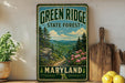 A vintage-style metal sign for the Green Ridge State Forest in Maryland, featuring a scenic mountain landscape with pine trees, flowers, and a winding path.