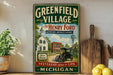A vintage-style sign for "Greenfield Village" featuring an illustration of a historic building, a car, and a windmill, with the text "The Henry Ford Experience America's history" and " Yesterday comes to life Michigan".