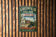 A vintage-style metal sign for the Greenfield Village, featuring a depiction of a historic building and a car, mounted on a wooden wall.