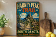 A vintage-style metal sign for the Harney Peak Trail in South Dakota, featuring a scenic mountain landscape with a trail sign and a sign indicating the trail's summit.