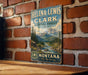 A vintage-style metal sign with a mountain landscape and a river, mounted on a brick wall.