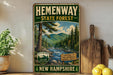 A vintage-style metal sign for Hemenway State Forest in New Hampshire, featuring a scenic image of pine trees, a river, and mountains.