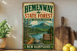 A vintage-style metal sign for Hemenway State Forest in New Hampshire, featuring a scenic mountain landscape and a trail sign.