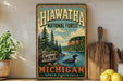 A vintage-style metal sign advertising the Hiawatha National Forest in Michigan, featuring a scenic image of a lake surrounded by pine trees and mountains.