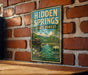 A vintage-style metal sign for Hidden Springs State Forest in Illinois, featuring a scenic landscape with a river and lush greenery.