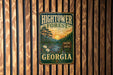 A vintage metal sign for the Hightower Forest in Georgia, featuring a scenic landscape with pine trees, a river, and a sunset.