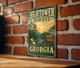 A vintage-style metal sign for the Hightower Forest in Georgia, featuring a scenic landscape with a river, trees, and a sunset.