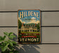 A vintage-style sign for Hildene, the Lincoln Family Home, is displayed on a wall next to a plant.