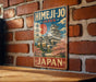 A vintage-style metal sign with the text "HIMEJI-JO JAPAN" and an illustration of a Japanese castle or pagoda surrounded by cherry blossom trees.