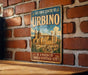 A vintage-style metal sign for the Historic Centre of Urbino, Italy, featuring an illustration of the historic center and the text "A UNESCO World Heritage Site".