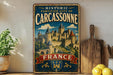 A vintage-style metal sign with the text "HISTORIC FORTIFIED CITY OF CARCASSONNE FRANCE" and an illustration of a castle, placed on a wooden surface next to a wooden cutting board and a potted plant.