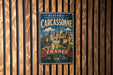 A vintage metal sign with the text "HISTORIC FORTIFIED CITY OF CARCASSONNE FRANCE" and an illustration of a castle, mounted on a wooden wall.