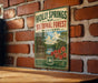 A vintage-style metal sign for the Holly Springs National Forest in Mississippi, featuring a scenic landscape with a lake, trees, and flowers.