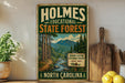 A vintage-style metal sign for Holmes State Forest in North Carolina, featuring a scenic illustration of a forest and mountains.