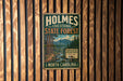 A vintage metal sign for Holmes State Forest in North Carolina, featuring a scenic image of a forest with a mountain backdrop.