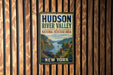 A vintage-style sign for the Hudson River Valley National Heritage Area in New York, featuring a scenic illustration of the river valley with mountains, buildings, and a train.