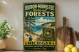 A vintage-style metal sign for Huron-Manistee National Forests, featuring a scenic landscape with pine trees, a lake, and mountains.