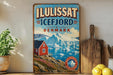 a vintage-style sign for Iluis SAT ICEFJORD DENMARK, with a scenic image of a red house on a cliff overlooking a body of water and mountains in the background.
