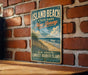 A vintage-style metal sign for Island Beach State Park in New Jersey, featuring a beach scene and the text "New Jersey's Longest Barrier Island" and "Beaches - Wild - Life - Adventure".