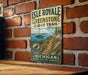 A vintage metal sign for Isle Royale Greenstone Ridge Trail, featuring a scenic mountain landscape with a moose and a path leading to the island.