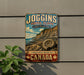 A vintage-style metal sign advertising the Joggins Fossil Cliffs, a world heritage site in Canada, with a scenic image of the cliffs and a spiral fossil.