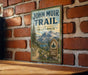 A vintage metal sign for the John Muir Trail in California, featuring a scenic mountain landscape and a trail marker.