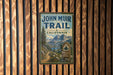 A vintage metal sign for the John Muir Trail in California, featuring a scenic mountain landscape and a trail marker.