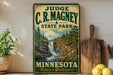 A vintage-style sign for C.R. Magney State Park in Minnesota, featuring a scenic illustration of a waterfall surrounded by pine trees.