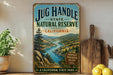 A vintage-style metal sign for the Jug Handle State Natural Reserve in California, featuring a scenic landscape with a river and mountains.