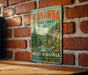 A vintage-style metal sign for Kanawha State Forest in West Virginia, featuring a scenic illustration of a forest landscape with a river and waterfall.