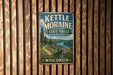 A vintage metal sign for Kettle Moraine State Forest in Wisconsin, featuring a scenic landscape with a river, mountains, and pine trees.