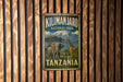 A vintage-style metal sign for Kilimanjaro National Park in Tanzania, featuring an illustration of a giraffe and an elephant in a natural landscape.