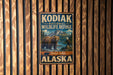 A metal sign with the text "KODIAK NATIONAL WILDLIFE REFUGE" and an illustration of a bear and eagle, mounted on a wooden fence.