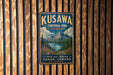 A vintage-style metal sign for Kuskawa Territorial Park in Yukon, Canada, featuring a mountain landscape and a lake.