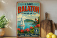 A vintage-style metal sign advertising Lake Balaton, Hungary, featuring a sailboat on the lake and a castle in the background.