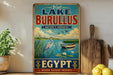 a vintage-style sign for Lake Burullus, Egypt, with a scenic illustration of a lake, a heron, and a boat. The sign encourages visitors to "Visit Lake Burullus" and highlights the "Nature's Paradise" of the region.