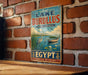 a vintage-style metal sign with a scenic illustration of a lake, a boat, and a bird, mounted on a brick wall.
