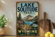 A vintage-style sign for the Lake Solitude Trail in Wyoming, featuring a scenic mountain landscape with a lake and pine trees.