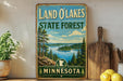 A vintage-style sign for the Land O'Lakes State Forest in Minnesota, featuring a scenic landscape with pine trees, a lake, and mountains.